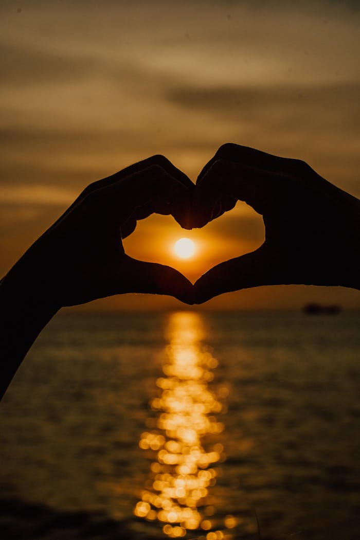 Silhouetted hands form a heart shape against a stunning ocean sunset backdrop.